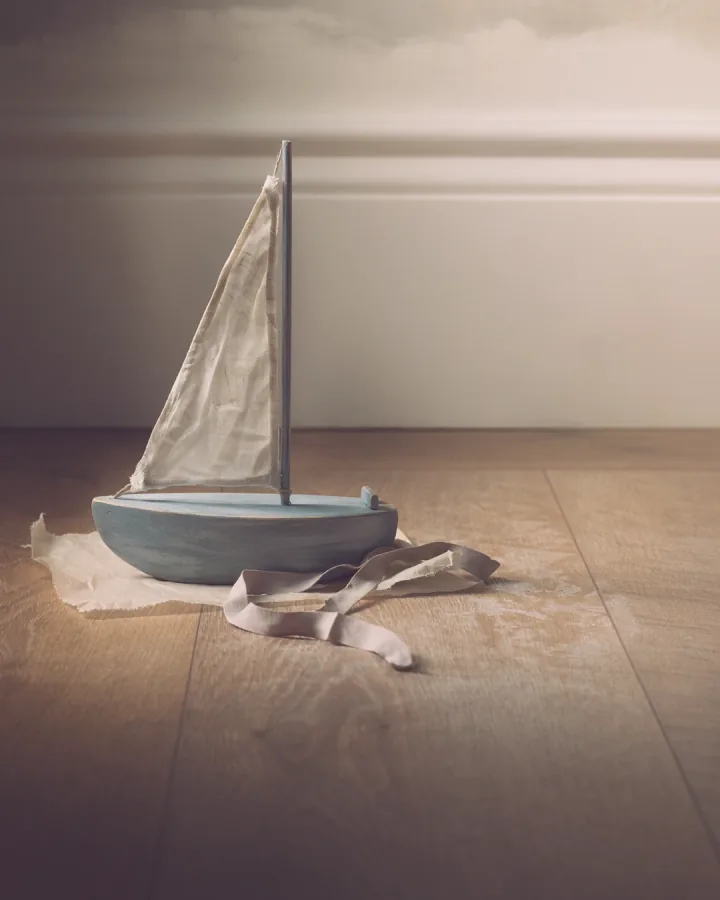 A painted wooden toy sailing boat on tissue on a bare wooden floor.