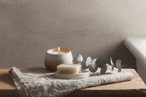 A folded towel, a bar of soap and a lit candle on a low stool beside a bath.