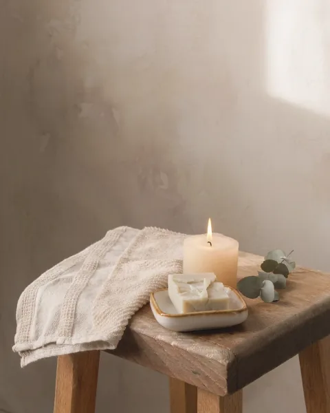 A folded towel, a bar of soap and a lit candle on a low stool beside a bath.