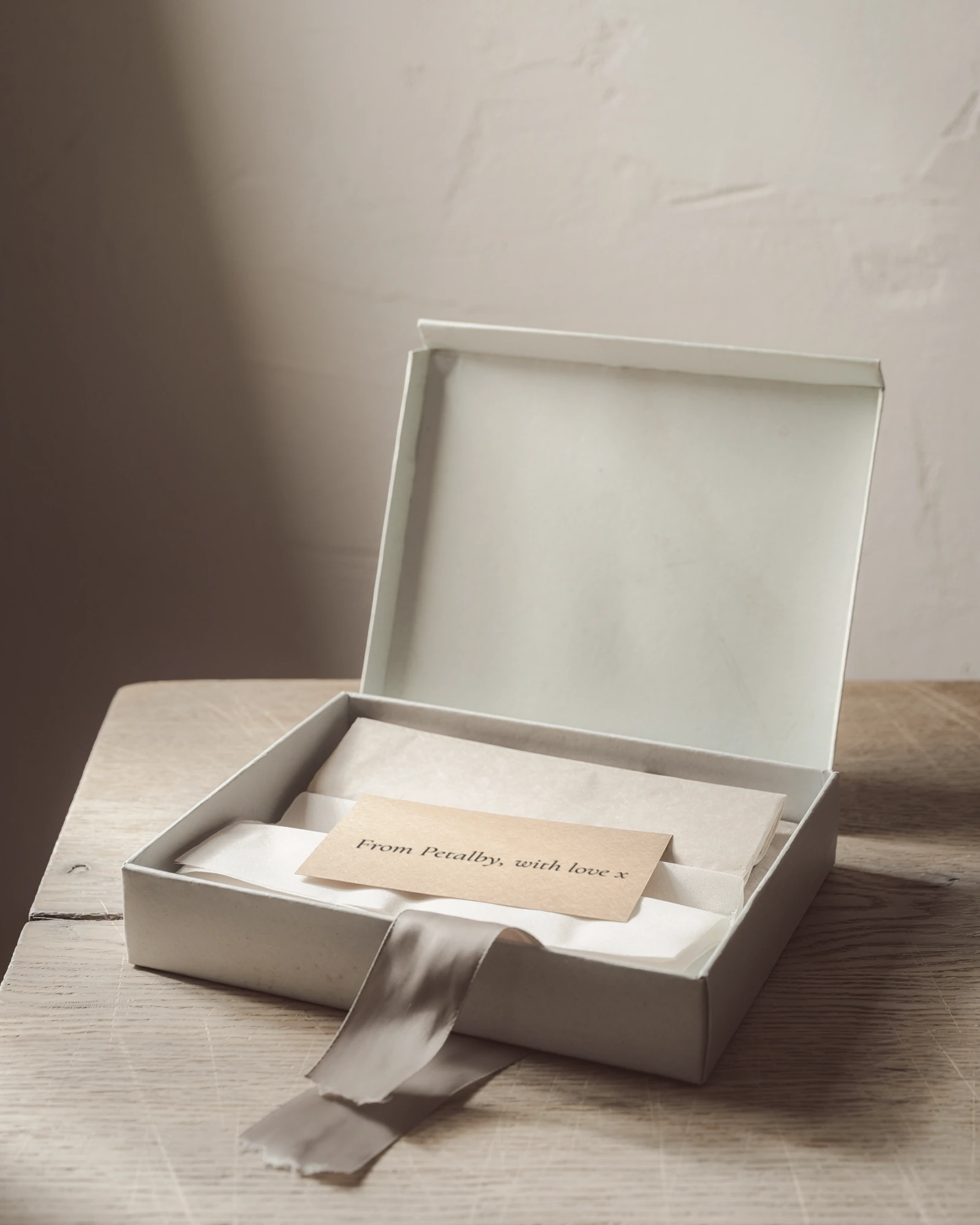 An open box with its lid raised, folded tissue inside, and a card that reads From Petalby, with love x, with a loose ribbon trailing across an oak table.