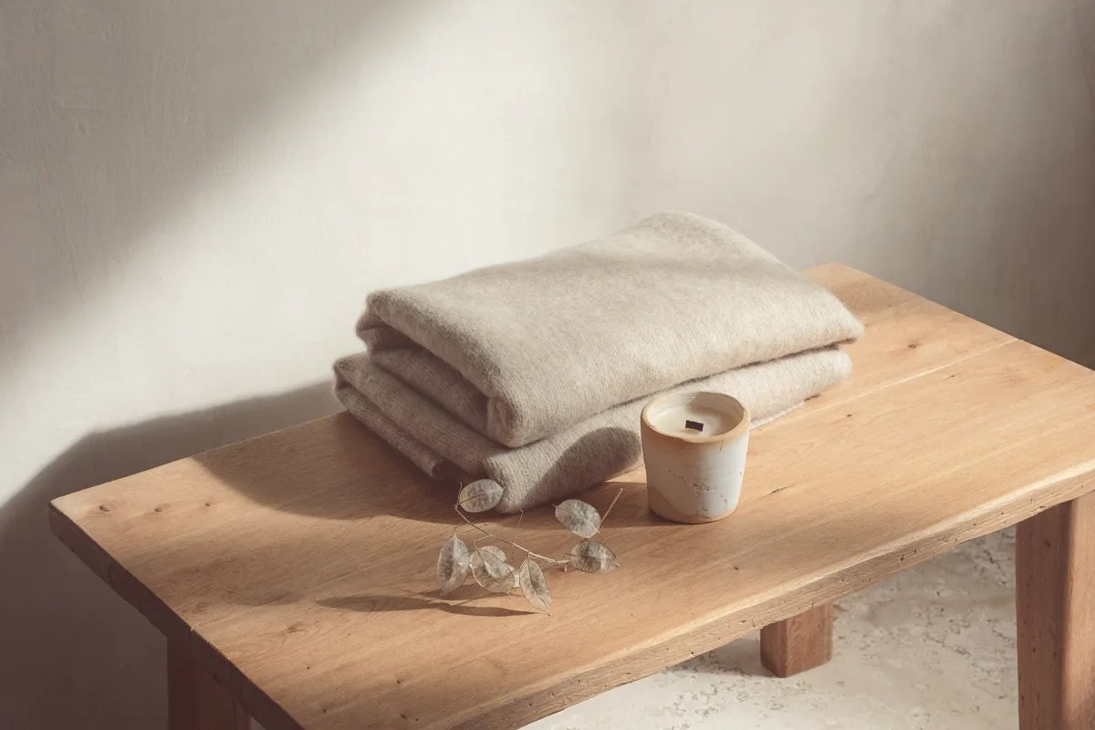 A folded wool blanket and a candle on a low table in soft shadow.