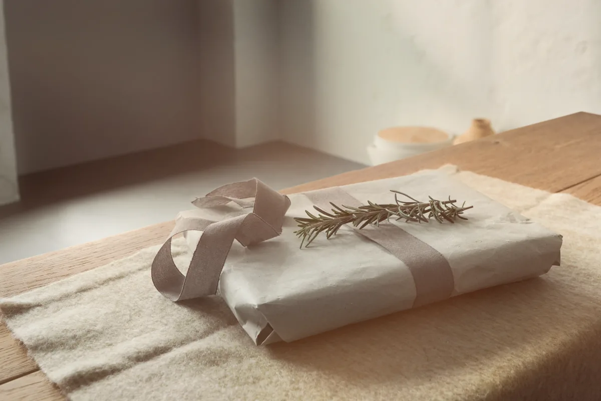 A paper parcel with a wide ribbon laid over it and a sprig of rosemary on a wool cloth