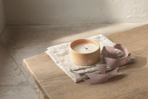 A candle in warm-white ceramic and a bar of hand-cut soap on a ceramic dish, with tissue and a loose ribbon on an oak table.