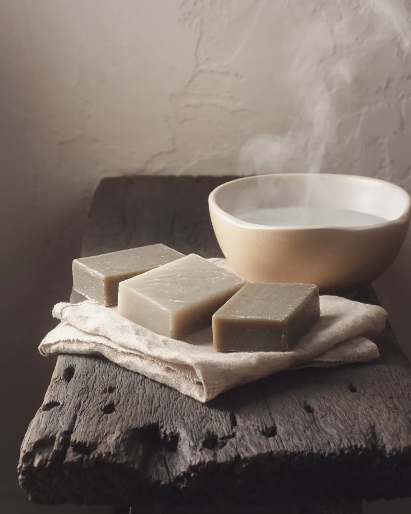 Three hand-cut bars of soap laid side by side on a linen cloth, beside a shallow ceramic bowl on a weathered oak block