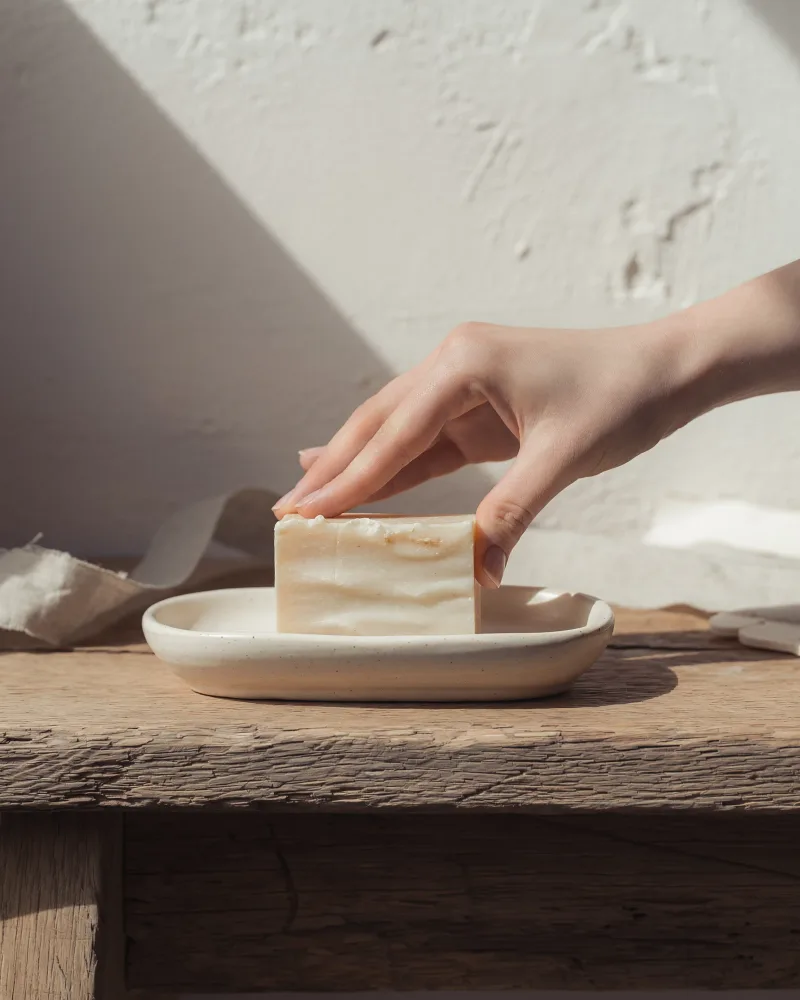 A hand resting on a hand-cut bar of soap on a shallow ceramic dish, on a worn oak bench.